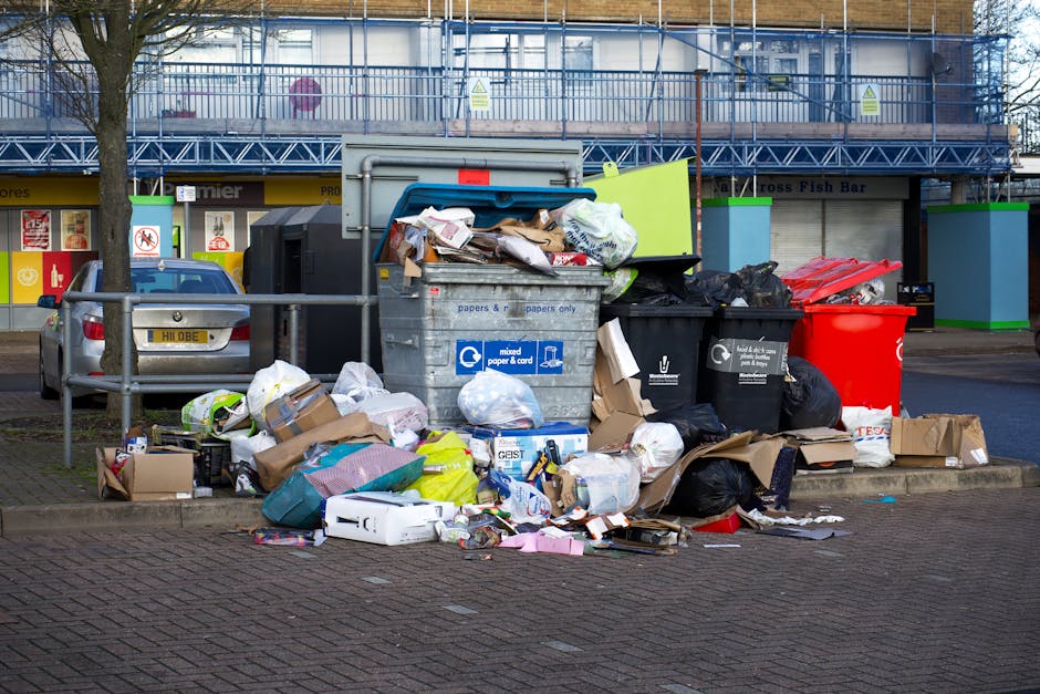 An outdoor scene showing several overflowing waste bins and scattered rubbish on a paved area in South Norwood. The waste includes cardboard boxes, plastic bags, paper, and packaging materials piled on the ground and around the bins. One larger recycling bin labeled for mixed paper and card is partially open with additional rubbish spilling out. There are black, green, and red bins, some with lids open, positioned near a railing and a parked silver car with a visible yellow license plate. In the background, there is a commercial building under construction or renovation with scaffolding and safety barriers, and a row of shops with signage. The scene is illuminated by natural daylight, indicating daytime, and the image relates to waste disposal laws and rubbish management in South Norwood, relevant to the services offered by Man with Van South Norwood for house removals and home relocation logistics.
