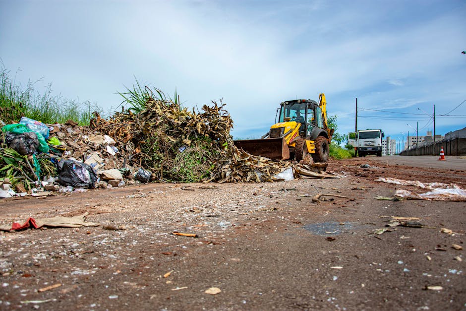 An outdoor scene showing several overflowing waste bins and scattered rubbish on a paved area in South Norwood. The waste includes cardboard boxes, plastic bags, paper, and packaging materials piled on the ground and around the bins. One larger recycling bin labeled for mixed paper and card is partially open with additional rubbish spilling out. There are black, green, and red bins, some with lids open, positioned near a railing and a parked silver car with a visible yellow license plate. In the background, there is a commercial building under construction or renovation with scaffolding and safety barriers, and a row of shops with signage. The scene is illuminated by natural daylight, indicating daytime, and the image relates to waste disposal laws and rubbish management in South Norwood, relevant to the services offered by Man with Van South Norwood for house removals and home relocation logistics.