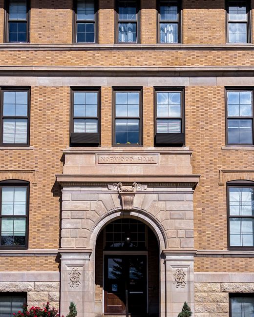 The image shows the exterior facade of Norwood Hall, a brick-built building with several evenly spaced windows featuring black window boxes. The entrance is centered and framed by a stone archway with