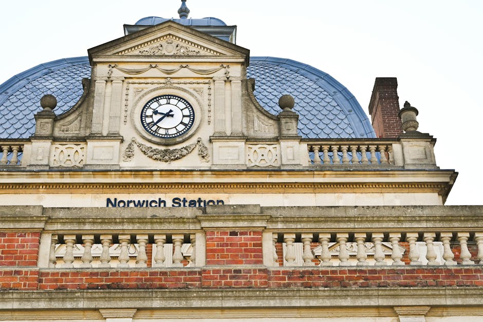 Close-up view of the upper part of Norwich Station's historic building facade, featuring ornate stonework, a large round clock with black Roman numerals and black hands displaying approximately 1:11, and a decorative garland beneath the clock. The building’s exterior includes beige stone and red brickwork, with a blue, crenellated mansard roof visible in the background. Additionally, a chimney with a brick base and chimney pipe is seen on the right side of the roof. The station name 'Norwich Station' is partially visible below the decorative stone elements. This image illustrates the exterior environment of a historic transportation hub, relevant to the context of home relocation or furniture transport in relation to [PAGE_TITLE], emphasizing the setting where professional removals services, such as those provided by [COMPANY_NAME], might operate during moving and packing processes.