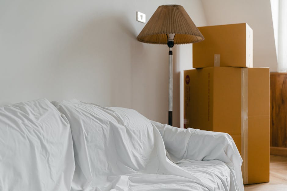 Indoor scene showing a living room corner with a white sofa covered by a protective sheet, positioned against a light-colored wall with a standard power socket visible near the ceiling. To the right, there are three large cardboard moving boxes stacked on top of each other, situated on the wooden floor close to a brown wooden cabinet. Behind the boxes, a tall floor lamp with a brown pleated lampshade stands, partially illuminating the space. The boxes appear to be sealed with packing tape and are likely used for home relocation, while the plastic-wrapped sofa indicates preparations for packing and moving. The environment suggests an ongoing house removals process, with items staged for transport, as might be coordinated by a moving service such as Man with Van South Norwood, reflecting the logistics involved in furniture transport and packing during home relocations.