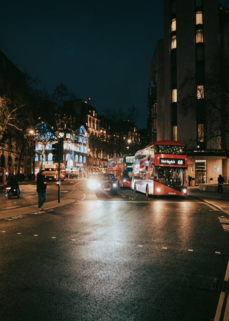 A nighttime city street scene shows a double-decker bus with the route number 68 and destination displayed on the front, waiting at a bus stop on the right side of the road. The street is illuminated by streetlights and building lighting, with a few pedestrians walking along the pavement. In the background, several parked cars and moving vehicles with headlights on are visible, along with illuminated windows in surrounding multi-story buildings. The road surface appears wet, reflecting the various light sources. On the left side, there is a crosswalk, while on the right side, a man is standing near the bus stop, possibly waiting to board or observing the traffic. The scene captures the typical urban environment of South Norwood at night, relevant to house removals, transport, and moving logistics, with an emphasis on the importance of planning for nighttime journeys during home relocations handled by companies like Man with Van South Norwood.