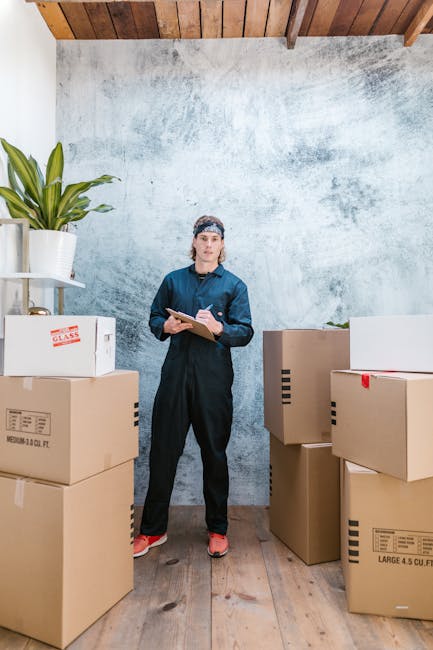 An indoor scene showing a woman in dark blue work overalls and a headscarf standing amidst several large cardboard moving boxes, some with red tape and labels. The boxes are placed on a wooden floor near a textured, light gray wall with a wooden ceiling above. To her left, there is a white shelving unit holding a green potted plant. The woman is holding a clipboard and pen, appearing to be checking or recording items as part of a home relocation or packing process. The environment suggests a preparatory stage for moving house, with the boxes ready for transport. The setting is well-lit, capturing the details of the packaging materials and furniture, consistent with the services offered by Man with Van South Norwood for removals and moving logistics.