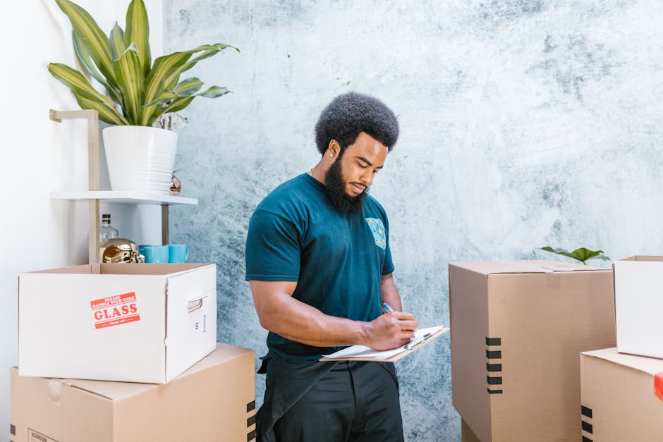 A man in a blue t-shirt and dark trousers is standing inside a room, surrounded by cardboard boxes, some labeled with red tape and others plain, used for house packing and moving. He is holding a clipboard and writing, with several boxes stacked around him, including one labeled ‘GLASS’ indicating fragile items. The room has a textured light-colored wall, and a white shelf with a large potted plant and decorative objects is visible in the background. This scene captures the packing and inventory process during a home relocation, with the man organizing and confirming items before transport. The environment suggests a professional moving or removals service, such as those offered by Man with Van South Norwood, assisting with furniture transport and packing for a house move in South Norwood (SE25). The lighting is natural, providing clear visibility for the detailed packing activity involving boxes, plants, and moving equipment.
