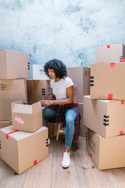 An indoor scene showing a woman in dark blue work overalls and a headscarf standing amidst several large cardboard moving boxes, some with red tape and labels. The boxes are placed on a wooden floor near a textured, light gray wall with a wooden ceiling above. To her left, there is a white shelving unit holding a green potted plant. The woman is holding a clipboard and pen, appearing to be checking or recording items as part of a home relocation or packing process. The environment suggests a preparatory stage for moving house, with the boxes ready for transport. The setting is well-lit, capturing the details of the packaging materials and furniture, consistent with the services offered by Man with Van South Norwood for removals and moving logistics.