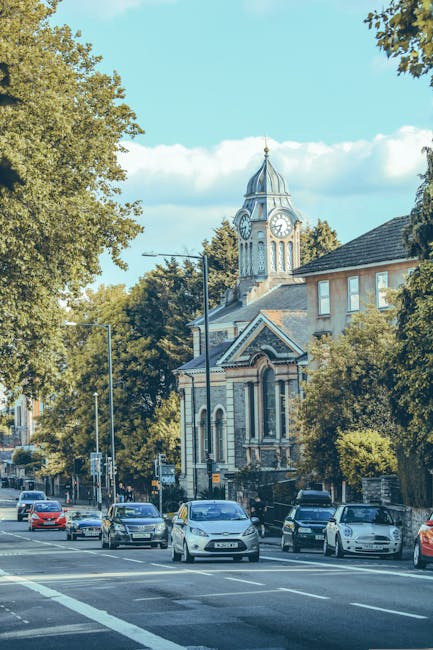 A street scene on Portland Road showing several cars parked along the curb and moving in both directions on a sunny day. Behind the vehicles, there is a historic church with a tall clock tower featuri