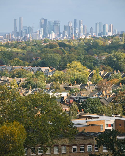 A wide aerial view of a residential neighbourhood with numerous houses featuring pitched roofs, surrounded by lush green trees in various shades of green and yellow, indicating autumn. In the background, a modern city skyline with tall glass skyscrapers and office buildings is visible under a partly cloudy sky. The trees form a transition zone between the suburban area and the urban centre. The image captures a peaceful, urban-to-rural landscape, suitable for illustrating home relocation or moving services in the context of house removals, as provided by Man with Van South Norwood. The natural lighting emphasizes the colours of the foliage and the cityscape, reflecting daytime conditions and the logistics involved in home packaging and furniture transport.
