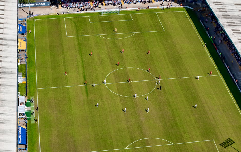 An aerial view of a football stadium pitch with players positioned across the field. The pitch is green with white lines marking the boundaries, center circle, and goal areas. Along the sidelines, there are several small structures and vehicles, possibly for team or staff use. The surrounding stands are filled with spectators, and the stadium's seating areas and entrances are visible. This image relates to the Selhurst Park football ground, where matchday arrangements or events may cause traffic or access considerations for house removals or home relocation services. Man with Van South Norwood offers support with furniture transport and packing during such busy periods, ensuring efficient moving logistics during matchdays or special events.