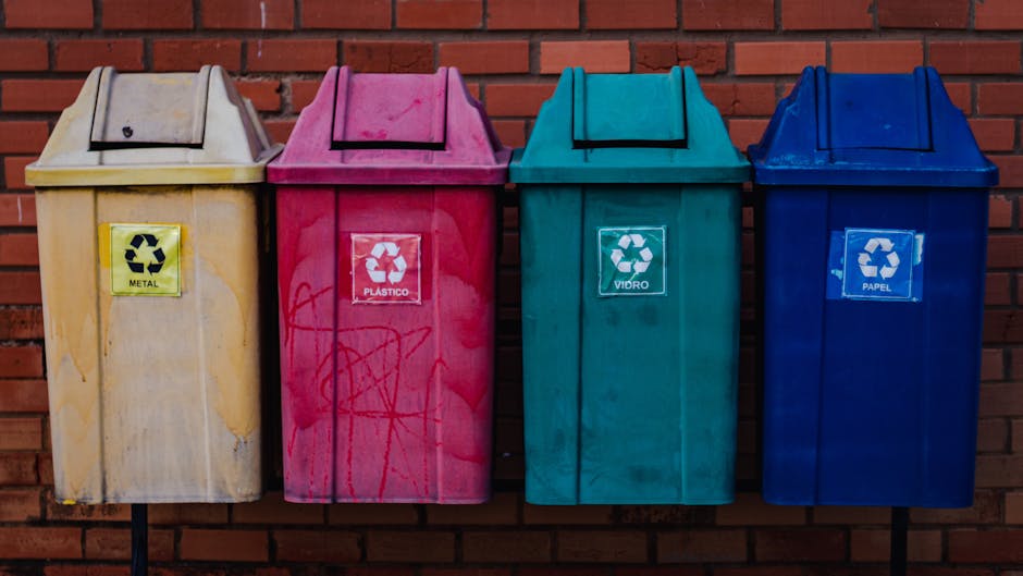 Four coloured recycling bins are positioned outdoors against a red brick wall, each designated for different waste types: a yellow bin for metal, a pink bin for plastics, a green bin for glass, and a blue bin for paper. The bins are made of plastic with hinged lids on top, and each has a printed recycling symbol alongside labels indicating the material category. The yellow metal bin shows some dirt and graffiti, while the others are clean and functional. They are placed on a paved surface, likely near a residential property or a communal area. In the context of house removals, these bins are typically used during packing and disposal stages, where proper waste segregation is essential in compliance with local regulations, such as those in Croydon. During a home relocation by Man with Van South Norwood, careful handling and disposal of waste materials like packaging and damaged items are part of the process, ensuring an efficient and environmentally responsible move.