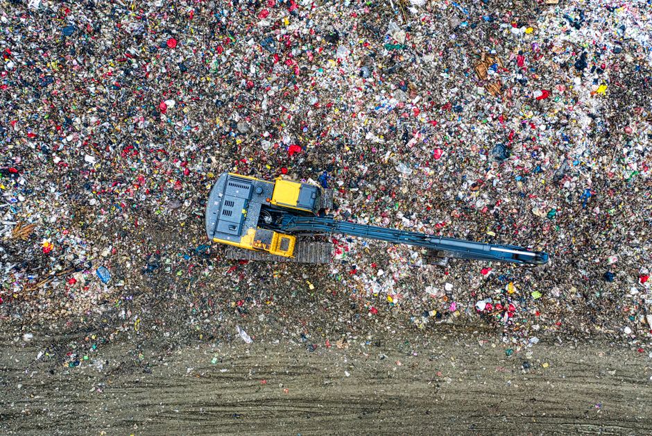 An aerial view of a yellow and black excavator with a long arm and bucket attachment, operating on a large area covered with mixed household waste including plastic, paper, packaging materials, and debris. The excavator is situated on a patch of bare, dark soil, with the waste scattered across the space, indicating waste collection or disposal activities related to house removals or clearing. The environment appears to be outdoors under natural lighting, with the equipment engaged in loading or sorting refuse, consistent with the context of waste disposal and packing materials management during a home relocation process, supported by Man with Van South Norwood.