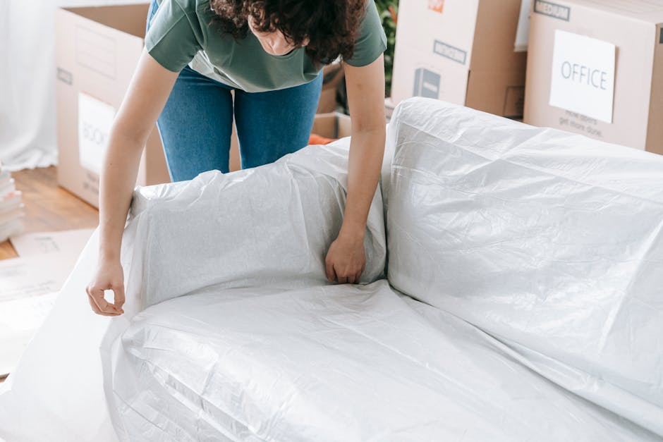 A person wearing a green top and blue jeans is in the process of wrapping and protecting a large sofa with white packing paper, likely preparing it for a house removal or furniture transport. The sofa is positioned inside a room, with the person carefully handling the wrapping material to ensure the furniture is secure for the moving process. In the background, there are cardboard boxes labeled for different rooms, such as 'OFFICE,' and some open packaging supplies on the floor, indicating a moving or packing operation. The setting appears well-lit with natural light coming into the room, and additional packing materials like cardboard and bubble wrap are visible, supporting the process of professional packing and moving, typical of services provided by [COMPANY_NAME], a company specializing in removals and home relocations.