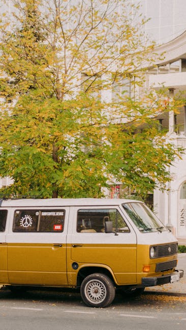 A mustard yellow vintage van parked on the pavement outside a residential property with white exterior walls and large windows. Behind the van, there is a large tree with yellow and green leaves, indicating autumn. The van appears to be used for home relocation or furniture transport, fitting within the context of removal services. The scene is well-lit with natural daylight, and the area looks calm and orderly. This image illustrates a typical setting for a house removals or moving company, such as Man with Van South Norwood, preparing for a local move along Station Road SE25, where careful loading and packing of belongings may take place inside or outside the property prior to transportation.