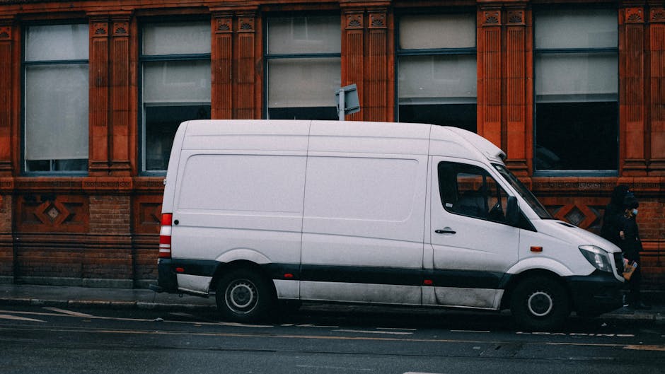 A white commercial van parked parallel to a sidewalk in front of a red brick building with decorative window surrounds. The van is positioned with the driver's side facing the curb, and it appears to be in the process of a home relocation or furniture transport, associated with moving and packing services offered by Man with Van South Norwood. The van's side door is closed, and the vehicle features black bump strips along the lower section. A person dressed in dark clothing is walking past the van on the pavement, which is also where moving boxes, wrapped furniture, and packing materials might typically be uploaded or unloaded during the loading process. The scene is captured in natural daylight, with the van occupying a significant portion of the street scene, emphasizing the logistical aspects of van access and parking for house removals in South Norwood.
