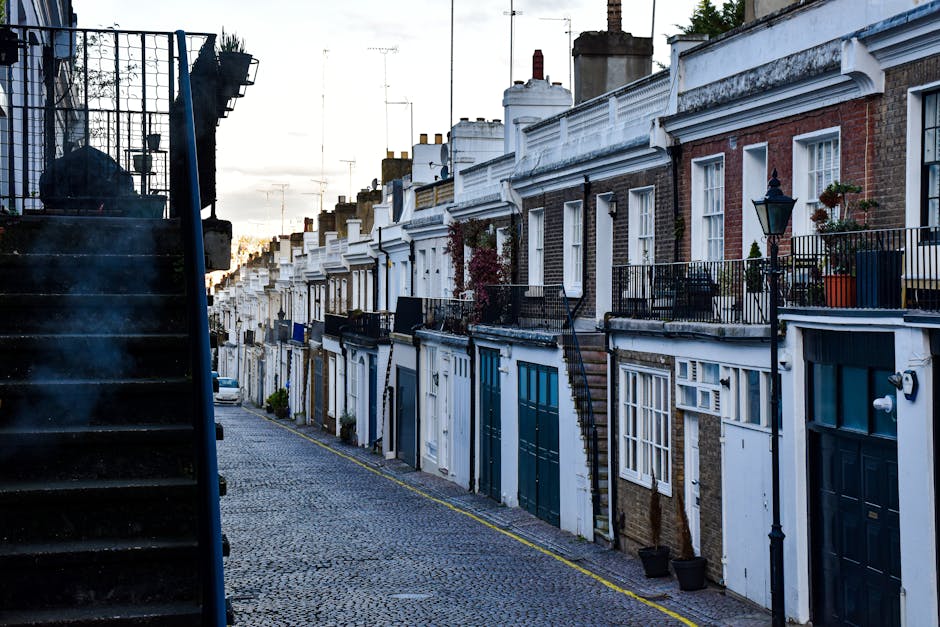A row of narrow, terraced houses on a street in Upper Norwood during daylight, with some houses featuring small balconies with black metal railings and potted plants, and others with white window frames and brick or painted facades. The street is paved with cobblestones, and a black external staircase is visible on the left side of the image. A parked car is seen further down the street, while the sky appears partly cloudy with hints of sunlight. This scene relates to home relocation and furniture transport, with [COMPANY_NAME] providing professional removal services in the area.