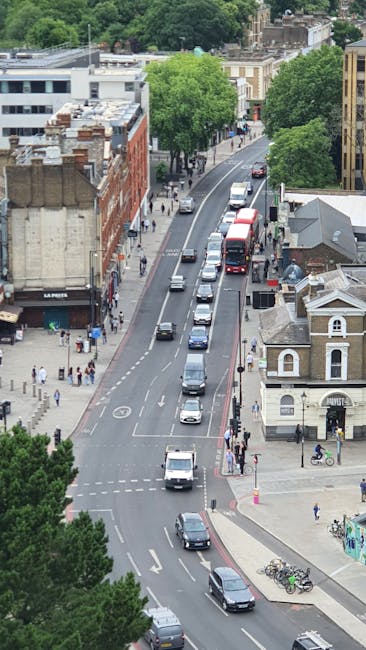 An aerial view of a busy urban street in South Norwood, showing a line of vehicles including cars, vans, and a double-decker bus moving along the road, with several pedestrians walking on the sidewalks. On the left side, there are commercial buildings, including a shop with a sign reading 'LA PIZZA,' and people standing near the storefronts. To the right, there are residential houses with small front yards, one of which has a bicycle parked outside. Tall, leafy trees line both sides of the street, contributing greenery to the scene. The street is managed with marked cycling lanes and pedestrian crossings, indicating an organized urban environment typical of local house removals and furniture transport activities. The lighting suggests it is daytime with overcast skies. This scene can be associated with the logistics of home relocation, packing, and moving services provided by Man with Van South Norwood, illustrating the busy street environment in which household furniture and boxes are transported during house moves.