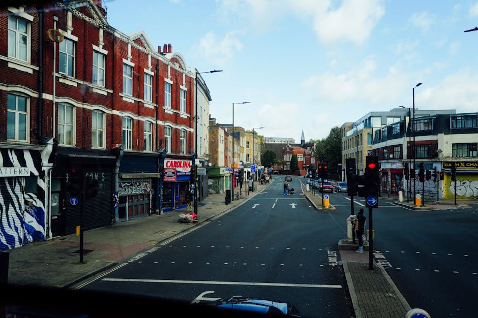 Photograph of a street scene in South Norwood during daylight hours, showing a row of mixed-use buildings with residential upper floors and ground-floor shops, including a barbershop and a cafe, with retail signage. The street is partially cordoned off with traffic lights showing red, and several parked cars are visible along the curb. On the pavement, cardboard boxes and packing materials are placed near the entrances of some shops, indicating recent or ongoing packing activities. A person is seen standing on the pavement on the right side of the image, possibly waiting or observing the scene. A large van with the branding of Man with Van South Norwood is parked on the street, close to a loading zone. The van’s rear doors are open, and furniture wrapped in protective blankets is being loaded or unloaded, with some items on dollies or trolleys. The environment is well-lit with natural daylight, and the sky above is partly cloudy. This scene exemplifies urban furniture transport and home relocation activities, with a focus on commercial streetscape and moving logistics.